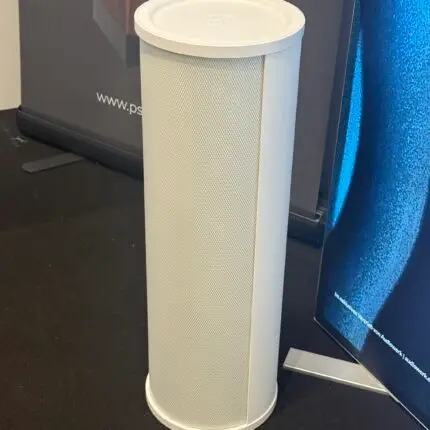 White cylindrical floor-standing speaker with a perforated grille on a black carpet, at an exhibition booth.